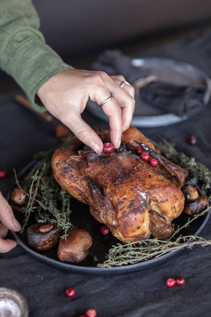 A close-up of a hand garnishing a perfectly roasted turkey with fresh cranberries, surrounded by herbs and mushrooms.